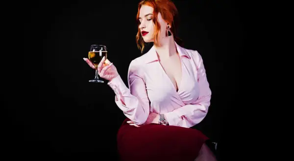 Donna con capelli rossi tiene un calice di vino da meditazione, con un'espressione pensierosa. Indossa una camicetta rosa e una gonna bordeaux.