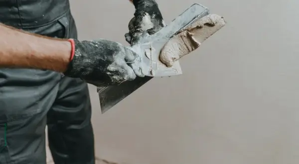 Ręce w rękawiczkach nakładają gładzie na ścianę za pomocą szpachli.