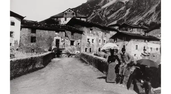 Un villaggio di montagna immortalato da Giovanni Verga fotografo. Coppie eleganti passeggiano con ombrelli, sullo sfondo case in pietra e pendii scoscesi.