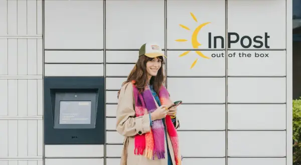Uśmiechnięta kobieta z telefonem stoi przy paczkomacie InPost. Wygodne akcje InPost to jej codzienność.