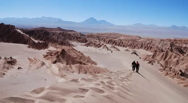 Dos excursionistas caminan por una duna en el desierto de Atacama, con formaciones rocosas y montañas al fondo bajo un cielo azul.
