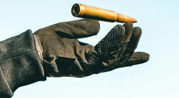 Una mano con guantes tácticos sostiene un cartucho dorado contra un cielo azul claro.
