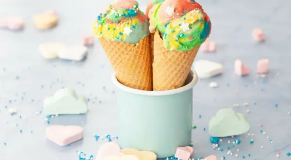 Zwei bunte Eiswaffeln mit Regenbogen-Eis und Streuseln stehen in einem hellblauen Becher. Marshmallows in Herz- und Wolkenform liegen verstreut.