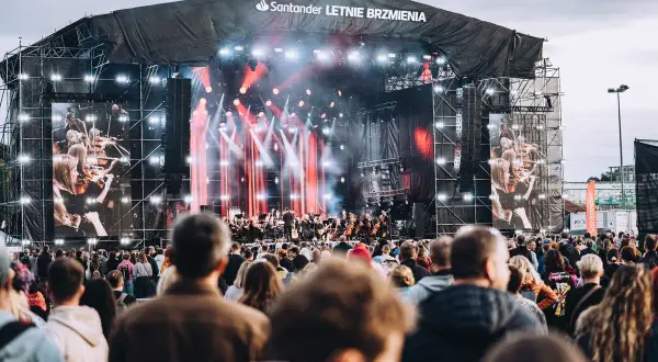 Koncert Santander Letnie Brzmienia z orkiestrą na żywo i ekranami pokazującymi muzyków. Tłum widzów czeka na występ.