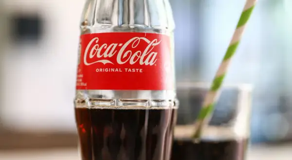 Szklana butelka Coca-Coli z czerwono-białą etykietą i słomką w tle.