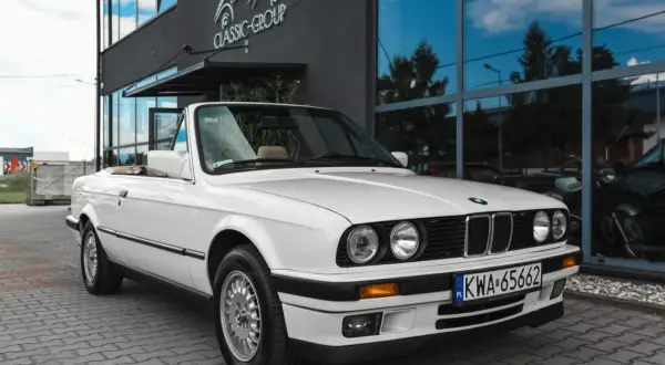 Białe bmw e30 cabrio z beżowym wnętrzem zaparkowane przed budynkiem z napisem "Classic Group".