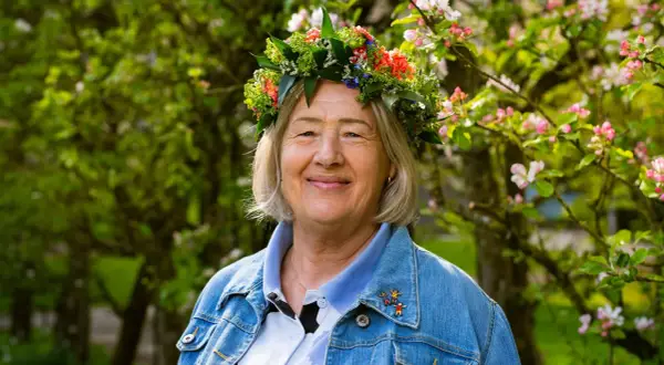 Glad kvinna med blomsterkrans, Bettan byvald, ler i solsken bland blommande träd.