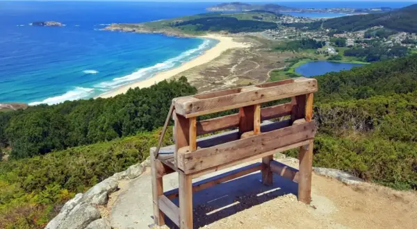 Banco de madera en un monte ventoso con vistas a una playa, el mar azul y un lago.