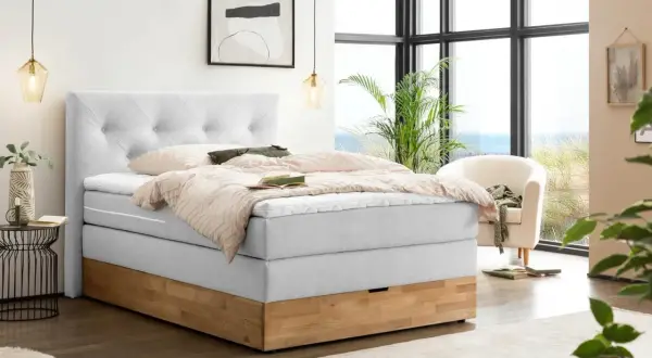 Ein hellgraues Boxspringbett mit Holzsockel und einem gesteppten Kopfteil. Die Nachteile eines Boxspringbetts, wie z.B. die Höhe, sind hier nicht ersichtlich.