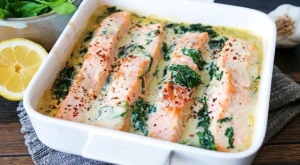 Vier saftige Lachsfilets in cremiger Spinatsauce, bestreut mit Gewürzen, in einer weißen Auflaufform. Daneben liegen frischer Spinat und eine Zitronenspalte.