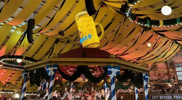Wielki, świecący kufel piwa wisi nad sceną z zespołem. Dekoracje w kolorach biało-niebieskim i żółtym tworzą atmosferę Oktoberfest 2025.