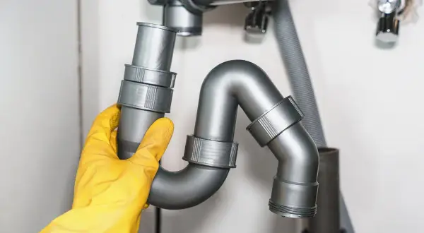 Main d'un plombier en gant jaune ajustant un siphon de rejet d'eau PVC sous un évier.