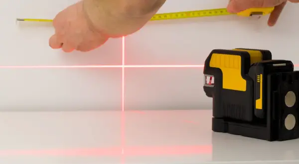 Poziomica laserowa emituje czerwone linie krzyżujące się na białej powierzchni, obok dłoni trzymającej miarkę.