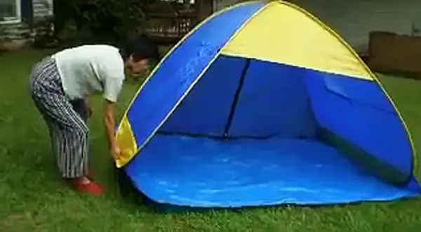 Osoba pochyla się nad rozkładanym namiotem plażowym w kolorze niebiesko-żółtym, stojącym na trawie.