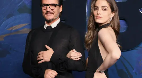 Pedro Pascal y una mujer elegante posan juntos. El actor, con gafas y un suéter negro, luce un cabestrillo. Ella, con un vestido negro y joyas brillantes, sonríe.