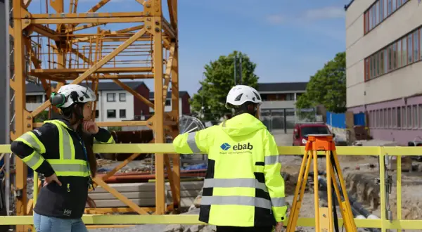 Två glada medarbetare i varselkläder och hjälmar på en byggarbetsplats. En stor kran syns i bakgrunden.