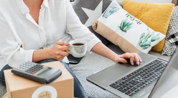 Kobieta z kawą uczy się, jak sprzedawać na OLX, korzystając z laptopa i kalkulatora.