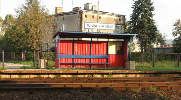 Przystanek kolejowy Warszawa Raków. Czerwona wiata z niebieską ławką i tablicą informacyjną. W tle budynek i wysokie drzewa.