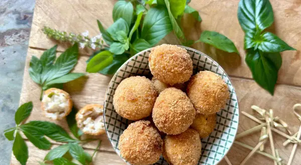 Goldbraune Arancini, frisch zubereitet nach einem tollen arancini rezept, liegen in einer Schale. Frische Kräuter und Holzspieße zieren die Holzplatte.