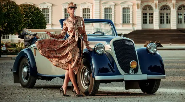 Elegancka kobieta w kwiecistej sukience retro z lat 20. pozuje przy zabytkowym kabriolecie przed pałacem.