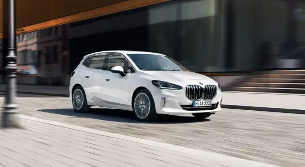 Biały BMW 2 Active Tourer w ruchu miejskim, z rozmytym tłem budynków i ulicy.