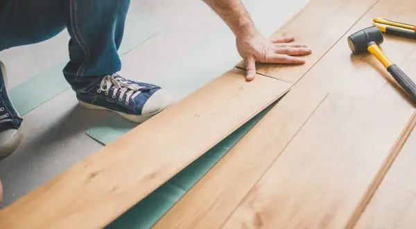 Pose d'un parquet flottant, une main ajuste une lame. Outil et maillet à proximité.