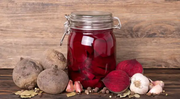 Ein Glas mit eingelegten, roten Bete Scheiben steht auf einem Holztisch. Daneben liegen frische rote Bete, Knoblauch und Gewürze. Das Bild zeigt den Prozess des rote Bete fermentieren.