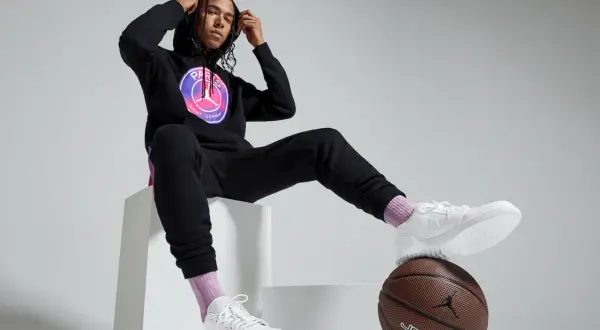 Mężczyzna w czarnej bluzie PSG Jordan z logo, czarnych spodniach dresowych, białych butach i różowych skarpetkach siedzi na białych sześcianach.