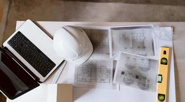 Na biurku leży laptop, biały kask, plany architektoniczne i poziomica. To kompletny zestaw do pracy nad projektem technicznym i prawem budowlanym.