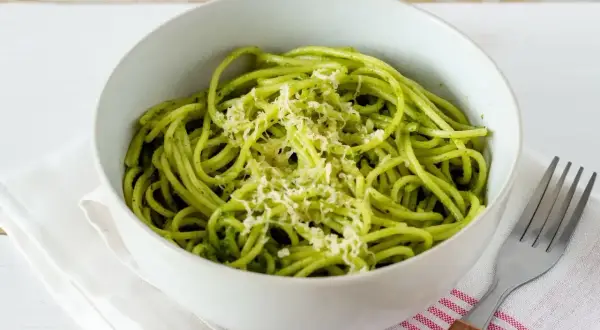 Miseczka spaghetti z zielonym, aromatycznym pesto z pokrzywy, posypana startym parmezanem. Obok leży widelec.