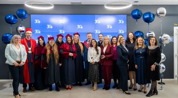 Grupa absolwentów i wykładowców świętuje 30-lecie swojej alma mater. Wszyscy ubrani są w togi i birety, a w tle wiszą balony.