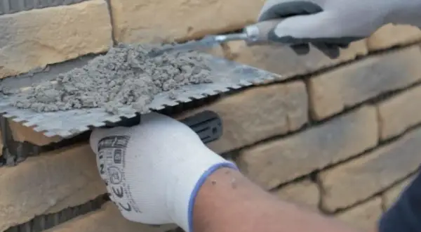 Ręce w rękawiczkach nakładają zaprawę na kamień dekoracyjny betonowy, przygotowując go do ułożenia na ścianie.
