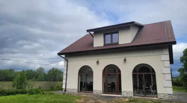 Nowy dom w Charsznicy z łukowymi oknami i brązowym dachem, otoczony zielenią i chmurami na niebie.