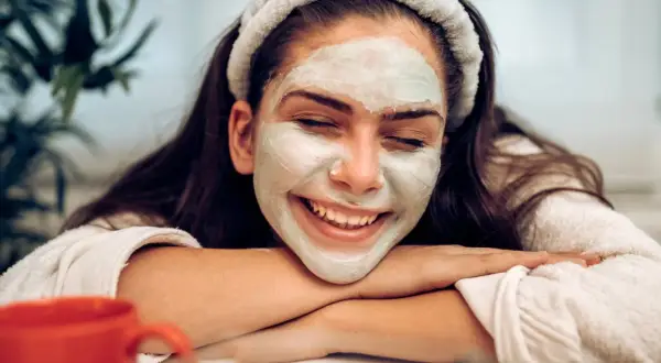 Lächelnde Frau mit Gesichtsmaske, die sich fragt: nach Gesichtsmaske eincremen oder nicht? Entspannter Moment mit rotem Becher.