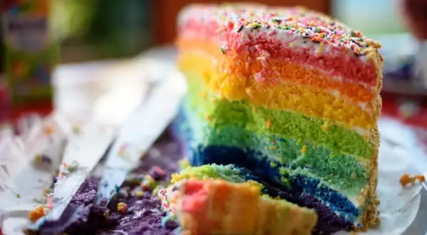 Ein Stück bunter **Regenbogen Kuchen** mit bunten Streuseln. Ein Stück ist bereits angeschnitten und liegt daneben.