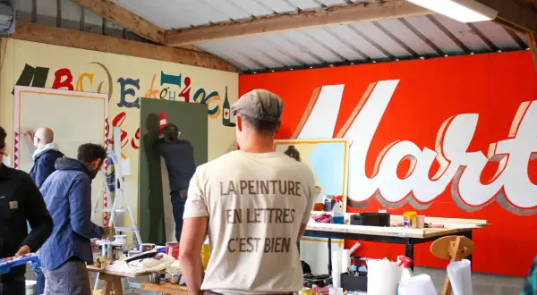 Des peintres travaillent dans un atelier, utilisant du matériel de peinture pour créer des lettres sur un mur. Le t-shirt d'un homme porte l'inscription "La peinture en lettres, c'est bien".