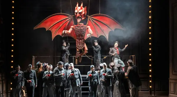 Bühnenszene der Oper Wuppertal: Eine Teufelsmarionette schwebt über Darstellern in Uniformen und Zylindern.