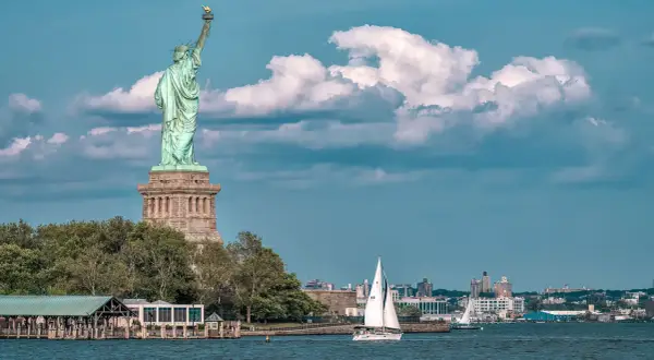 Statua Wolności na tle chmur, żaglówka na wodzie. Zastanawiasz się, ile kosztuje bilet na statek z Polski do USA?