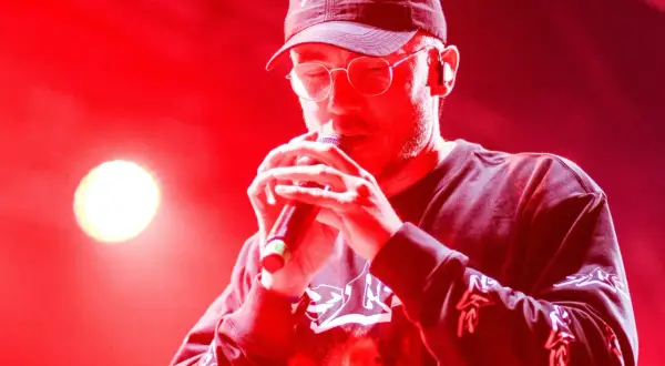Raper w czapce i okularach śpiewa do mikrofonu. Jego album zdobył Fryderyka w kategorii album roku (hip-hop).