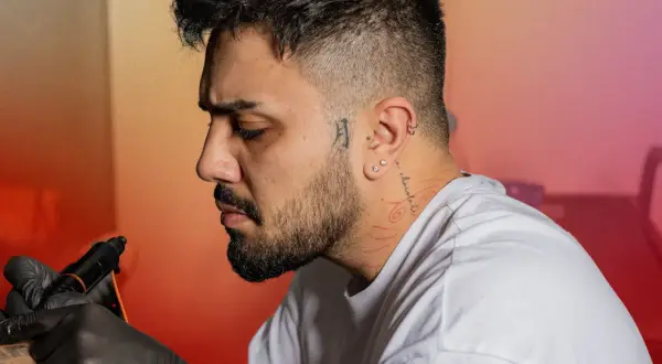 Un homme se fait tatouer le cou, un tatouage homme cou discret avec des motifs rouges. Il porte des gants noirs et une montre.
