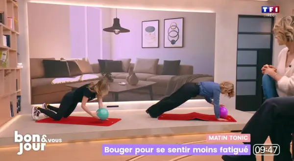 Deux femmes font des exercices sur des tapis rouges. L'une d'elles tient un ballon vert, l'autre un ballon violet. Elles semblent faire du sport tous les jours pour rester en forme.