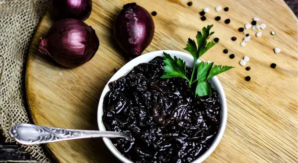 Schwarze, karamellisierte balsamico zwiebeln in einer Schüssel, garniert mit Petersilie. Daneben rote Zwiebeln und Pfefferkörner.