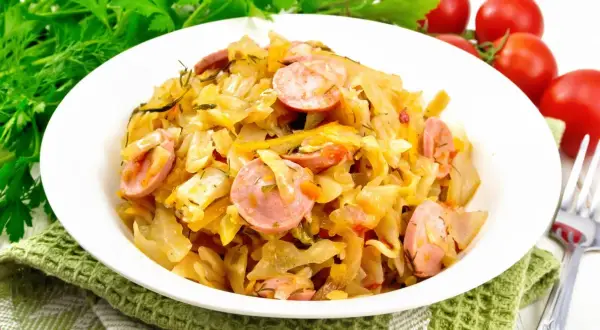 Pyszny bigos z młodej kapusty z kiełbasą, podany na białym talerzu, ozdobiony zieloną natką i pomidorkami.