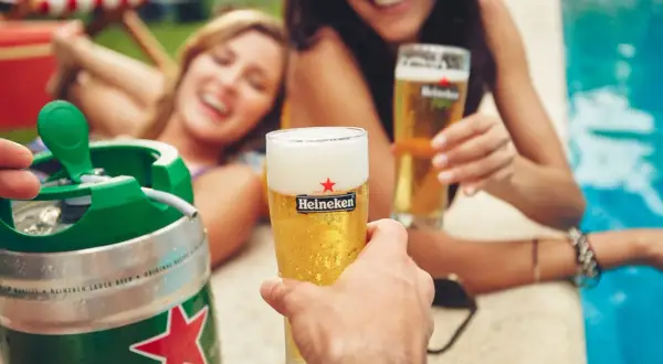 Un fusto di birra Heineken pronto per essere spillato, con due donne sorridenti che brindano con bicchieri di birra vicino a una piscina.