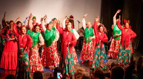 Un grupo de mujeres baila sevillanas de amor con trajes coloridos. Sus faldas amplias y mantones vibrantes llenan el escenario de alegría.