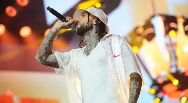 Malik Montana na scenie, przygotowuje się do swoich **koncertów 2025**. Wokół niego migoczą światła sceniczne.