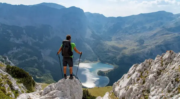 Trekking w Czarnogórze: turysta z plecakiem i kijkami stoi na skale, podziwiając górskie jezioro i pasma górskie.