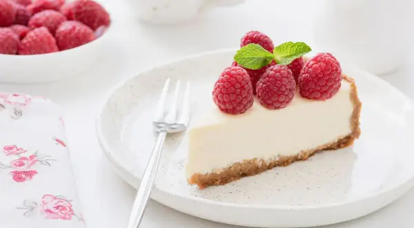 Una fetta di torta fredda allo yogurt, guarnita con lamponi freschi e menta, su un piatto con forchetta.