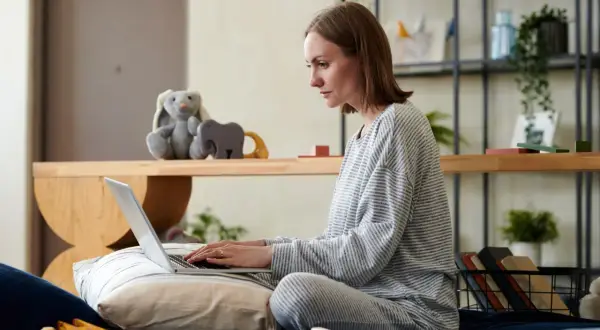 Kobieta w piżamie siedzi na łóżku, pisząc na laptopie. Obok pluszowy królik i zabawki. Przygotowuje wniosek o urlop rodzicielski.