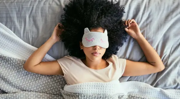 A woman with a large afro wears a cat-ear sleep mask, resting in bed. She's trying to figure out how long does it take to catch up on sleep.